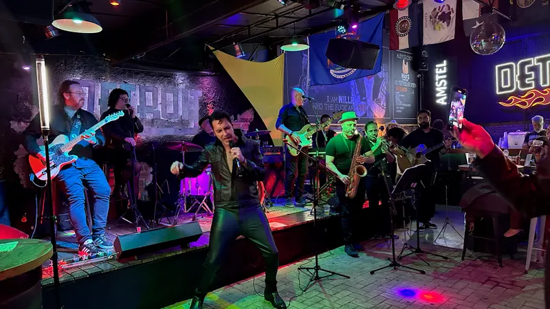 A banda completa no palco, com guitarras, baixo, sax e trompete, e Caio de couro preto à frente
