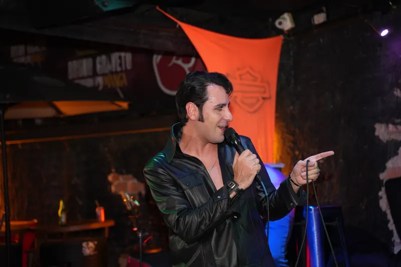 Caio de couro preto cantando ao microfone e apontando para a plateia, sorrindo