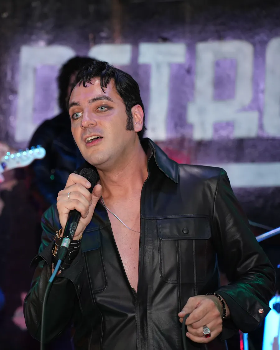 Caio Presley de jaqueta de couro preto cantando ao microfone, com o guitarrista ao fundo