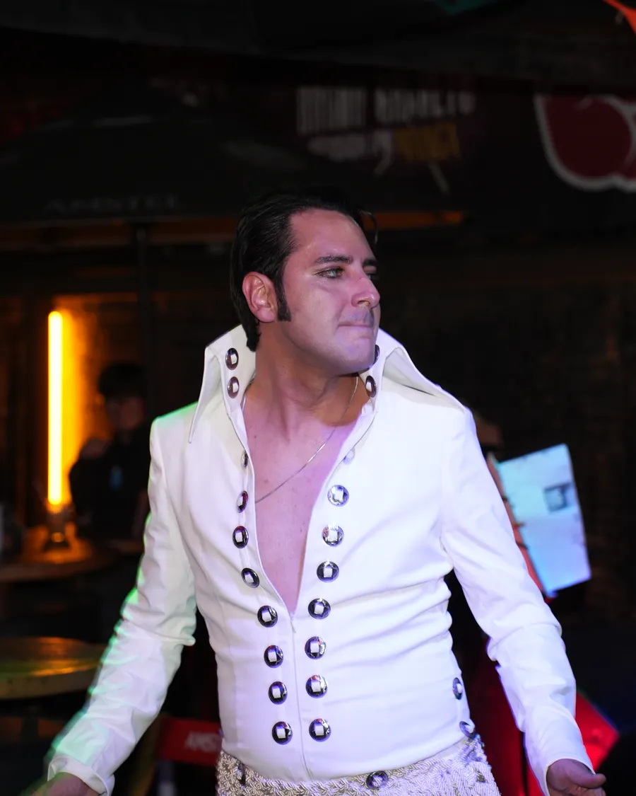Caio Presley de macacão branco com tachas prateadas e gola alta, de perfil, no meio de um passo de dança